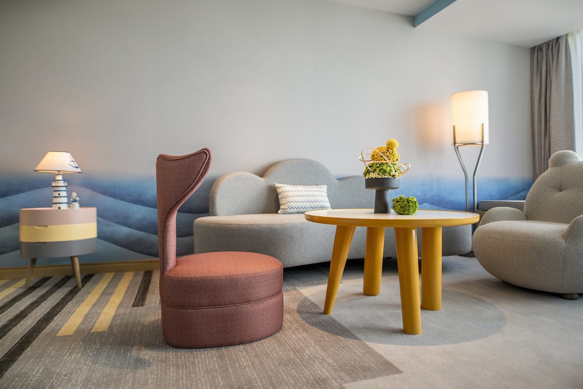 Une chambre d’hôtel moderne avec un canapé gris, un fauteuil rouge, une table ronde en bois et une lampe.