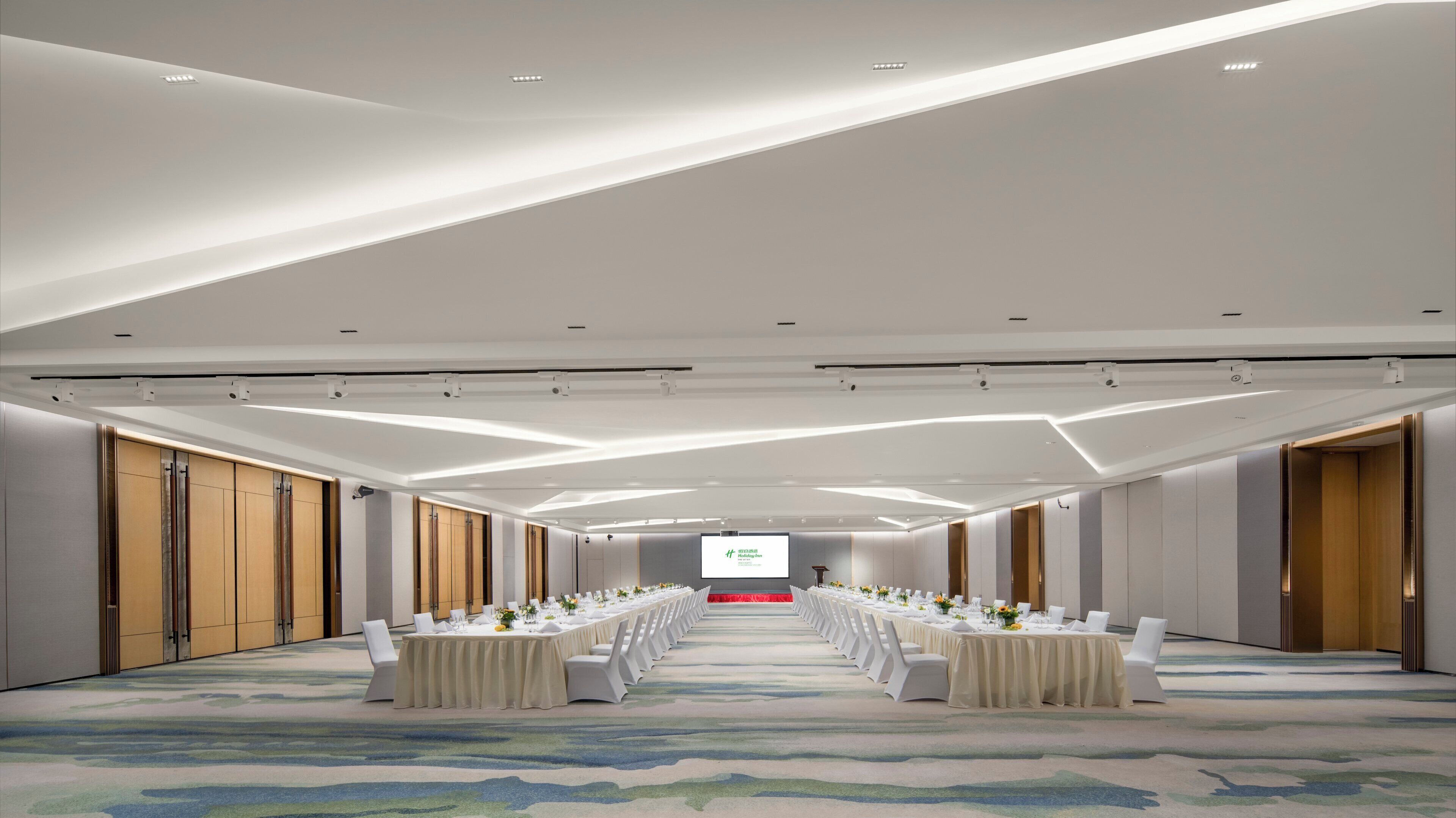 Un salón de conferencias con una larga mesa dispuesta para una reunión, equipado con una pantalla de proyector al frente.