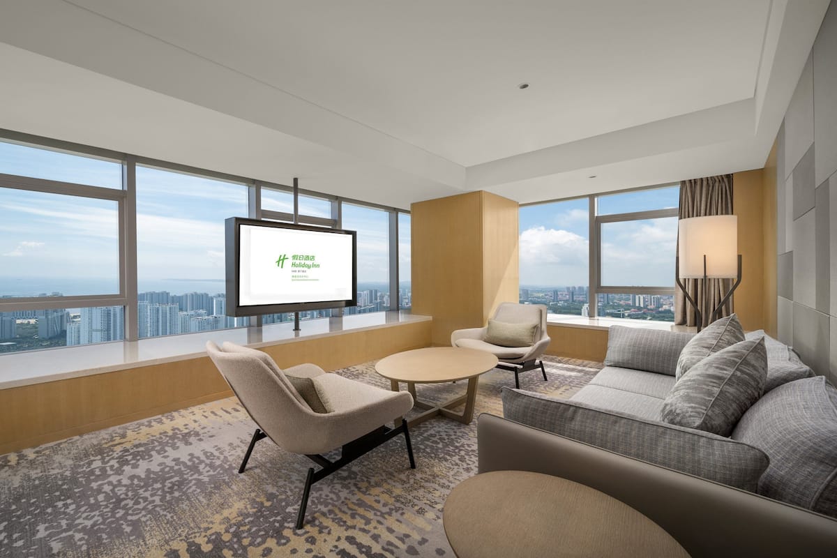 Une chambre d’hôtel moderne dotée d’une grande fenêtre donnant sur la ville, d’un téléviseur à écran plat, d’un canapé, d’un fauteuil et d’une petite table.