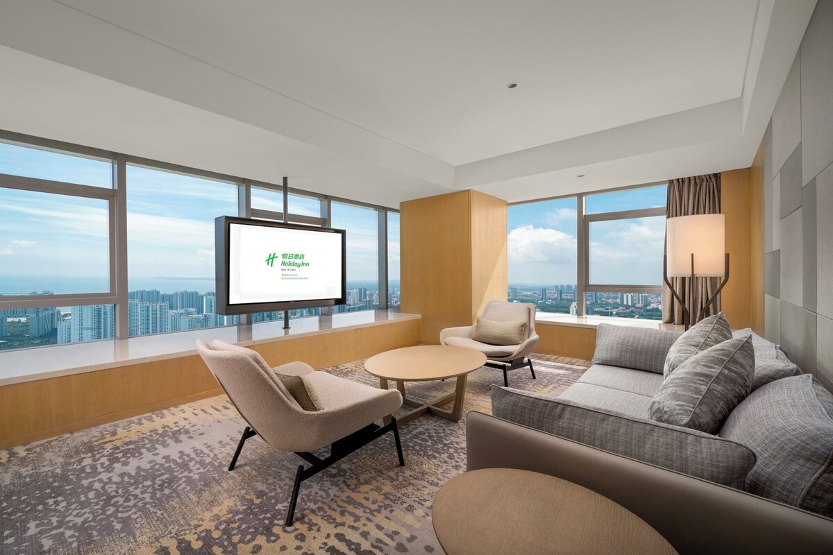 Une chambre d’hôtel moderne dotée d’une grande fenêtre donnant sur la ville, d’un téléviseur à écran plat, d’un canapé, d’un fauteuil et d’une petite table.