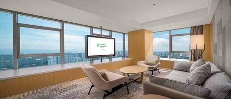 A modern hotel room with a large window offering a city view, a flat-screen TV, a sofa, a chair, and a small table.