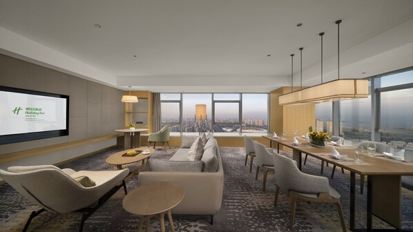 A modern hotel room with a large city view, a comfortable seating area, and a dining table set for two.