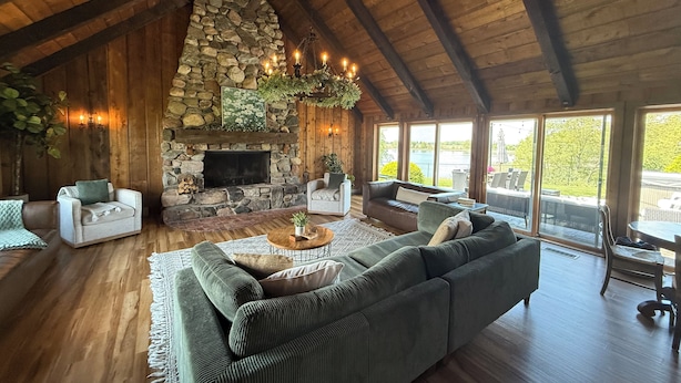 A cozy living room with a stone fireplace, wooden beams, and large windows overlooking a scenic outdoor area. - Secluded Lakefront Chalet  (Grass Lake)