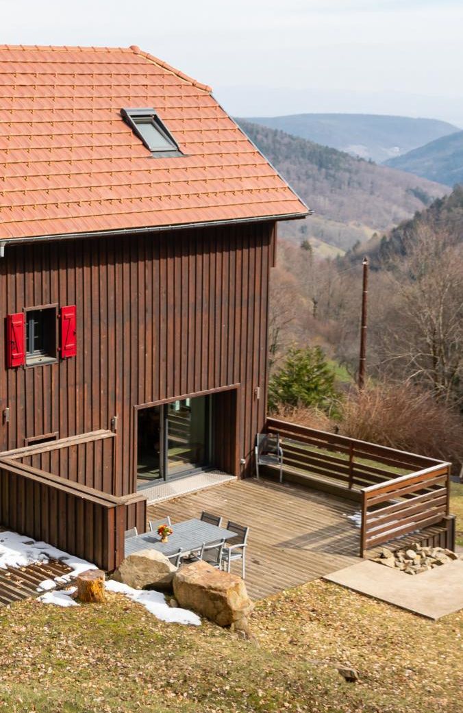 Un edificio de madera con techo inclinado, una ventana pequeña y una terraza con mobiliario exterior.