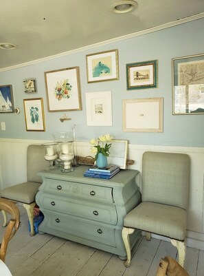 A room with a light blue wall, a wooden dresser with drawers, a chair, and various framed pictures. - Adorable Amagansett Cottage, Walk to Town or Beach (East Hampton)