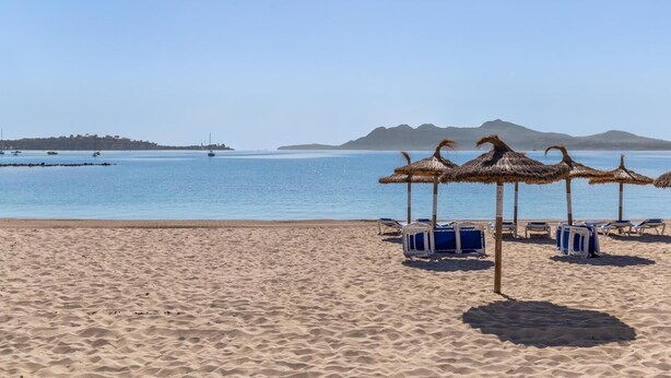 A beach with thatched umbrellas and sun loungers, a calm sea, and distant hills. - Beautiful, 3 bedroom, ground floor, sea front property, large furnished terrace (Puerto de Pollensa)