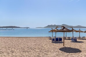 A beach with thatched umbrellas and sun loungers, a calm sea, and distant hills.