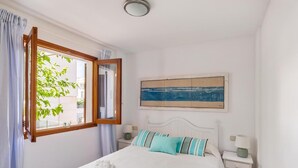 A bedroom with a bed, a nightstand, a lamp, a window with curtains, and a painting on the wall. - Beautiful, 3 bedroom, ground floor, sea front property, large furnished terrace (Puerto de Pollensa)