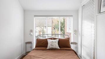 A bedroom with a bed, two bedside lamps, a window with blinds, and a view of buildings outside.