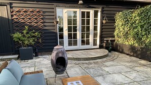 A patio with a built-in wood stove, a wooden table with a mug and a booklet, and a grey sofa. - Pet Friendly With Enclosed Leafy Outdoor Space (Saffron Walden)
