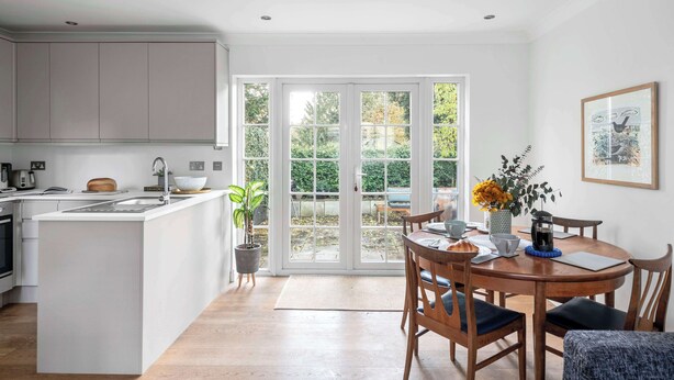 A modern kitchen with white cabinetry, a central island, and a dining area with wooden chairs and a table. - Pet Friendly With Enclosed Leafy Outdoor Space (Saffron Walden)