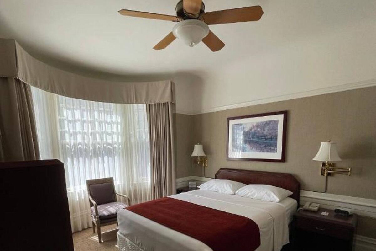 Une chambre d’hôtel comprenant un lit, une chaise, une table de chevet, une lampe, un ventilateur de plafond et une fenêtre avec des rideaux.