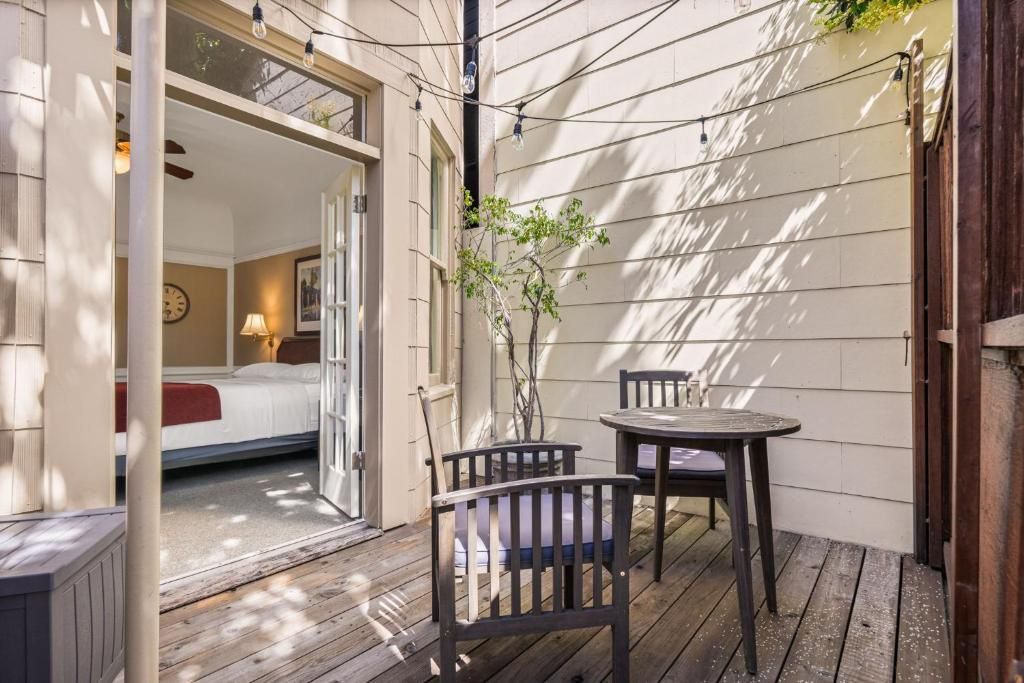 A cozy outdoor patio with a wooden deck, a small table, two chairs, and a view into a bedroom with a bed and a nightstand.