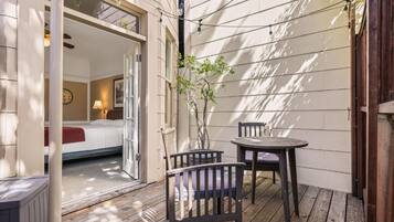 A cozy outdoor patio with a wooden deck, a small table, two chairs, and a view into a bedroom with a bed and a nightstand.