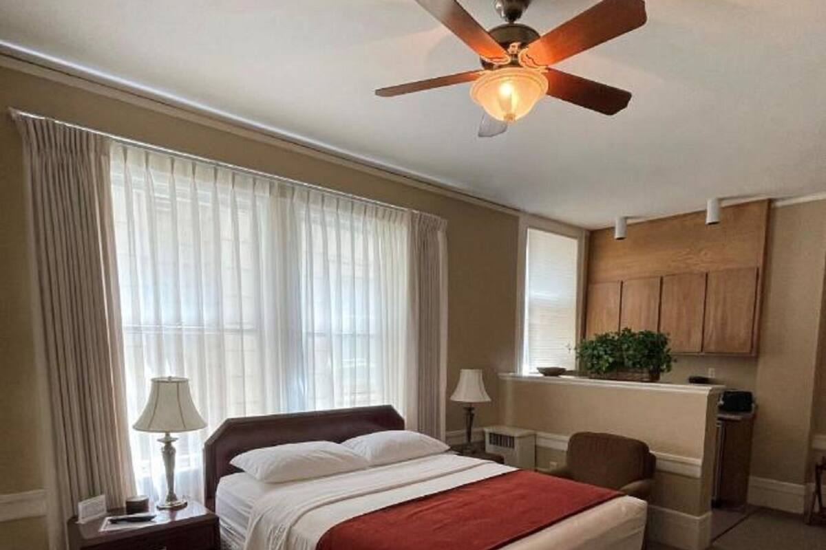 Une chambre d’hôtel équipée d’un lit, d’un bureau, d’une chaise, d’un ventilateur de plafond et d’une fenêtre avec des rideaux.
