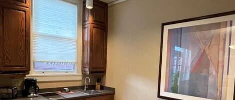 Una cocina con armarios de madera, horno empotrado, lavamanos y una ventana con persianas. Se observa una mesa de comedor con sillas y un cuadro en la pared.