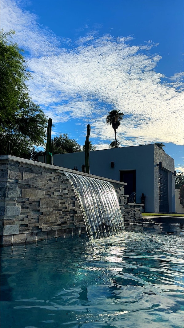 A swimming pool with a waterfall feature, a palm tree, and a building in the background.
