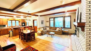 A spacious living area with a wooden floor, a fireplace, a dining area with a table and chairs, and a kitchen in the background.