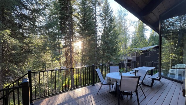 A wooden deck with outdoor furniture, surrounded by tall trees and a view of a cabin.