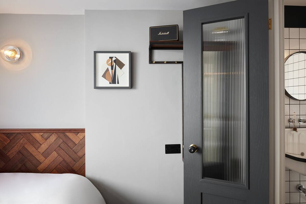 Habitación de hotel con cabecera de madera, una obra de arte abstracta enmarcada, una placa con nombre, una puerta de cristal satinado y una pared de azulejos.