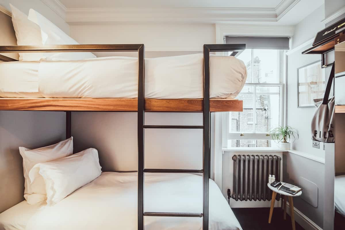 Dormitorio con literas, ropa de cama blanca, un radiador, una mesita con un libro y una ventana con vistas a edificios.