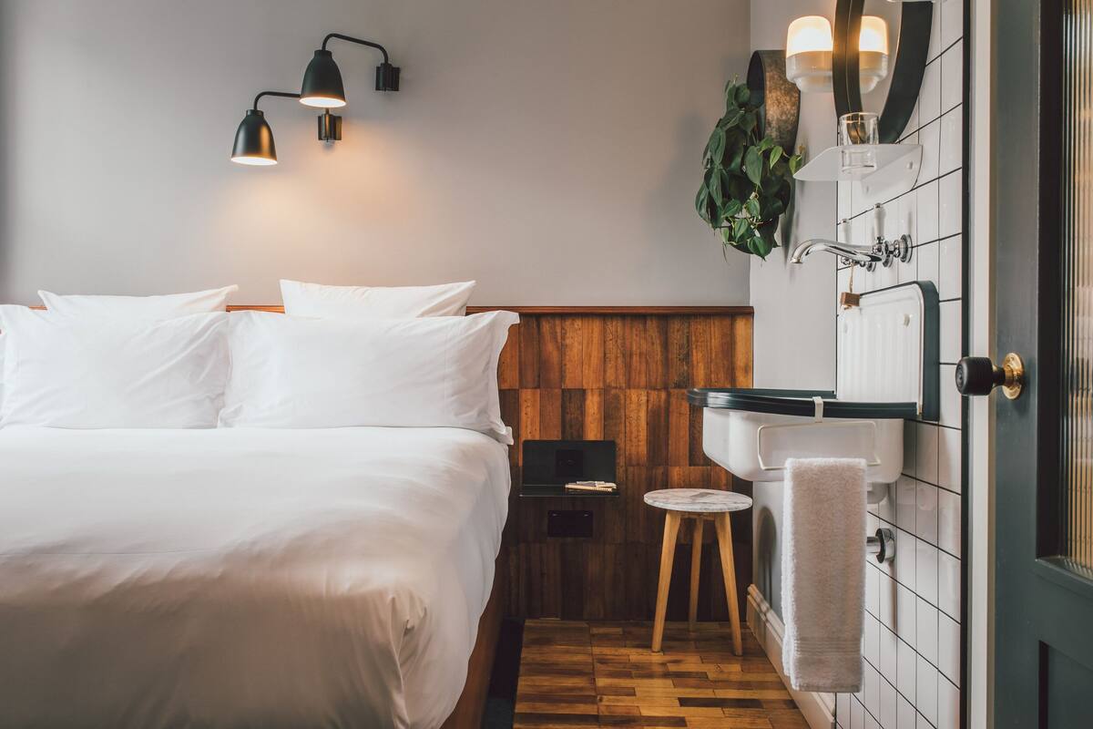 Una habitación de hotel con cabecera de madera, cama con sábanas blancas, una mesita de noche con lámpara, un espejo redondo y un inodoro empotrado con una toalla colgada al lado.