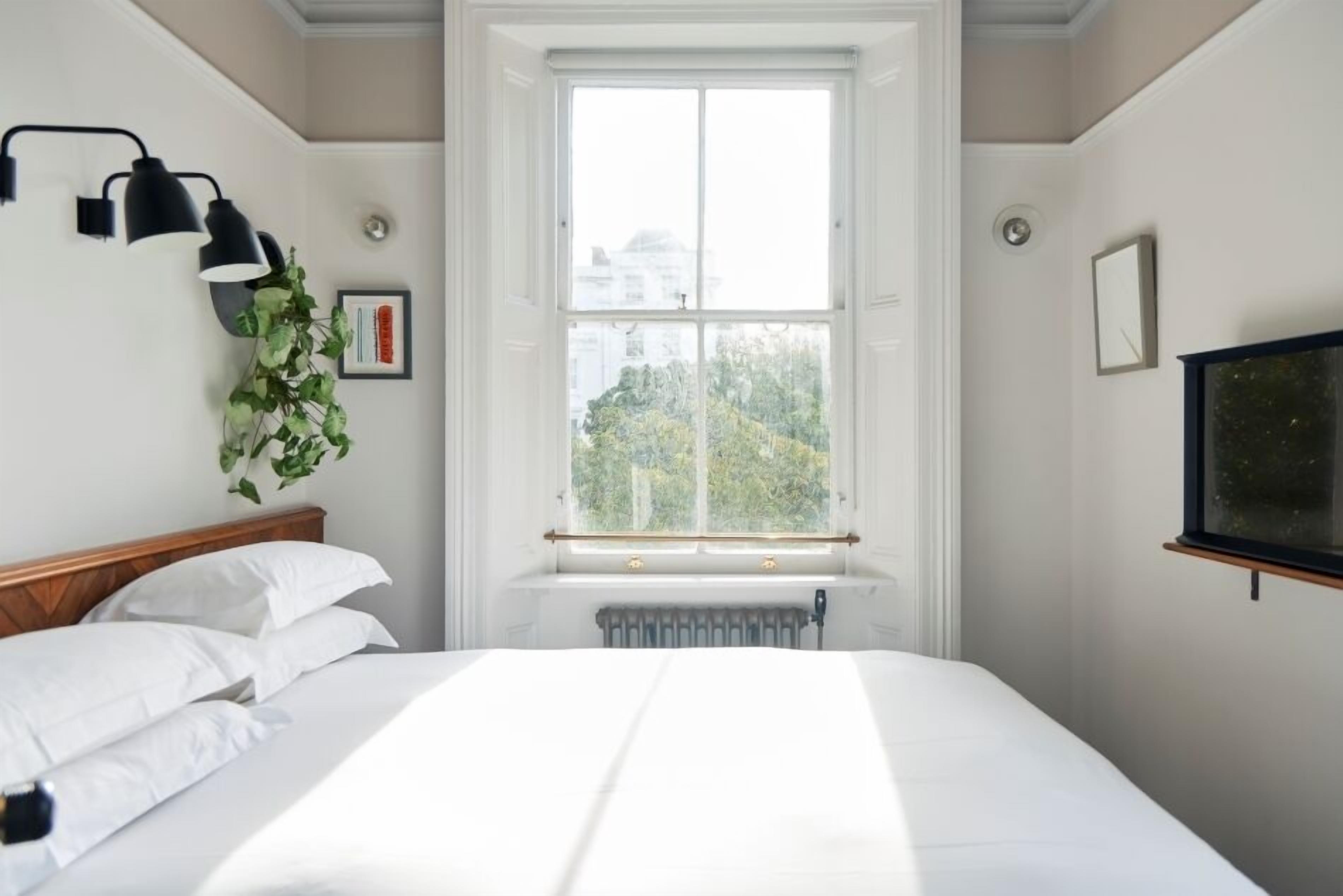 A bedroom with a large window, a radiator below it, a bed with white linens, a black wall-mounted lamp, and a framed picture on the wall.