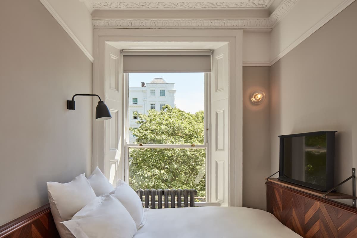 Un dormitorio con una cama, un radiador, un televisor sobre un soporte de madera y una ventana que ofrece vistas a un edificio y árboles.