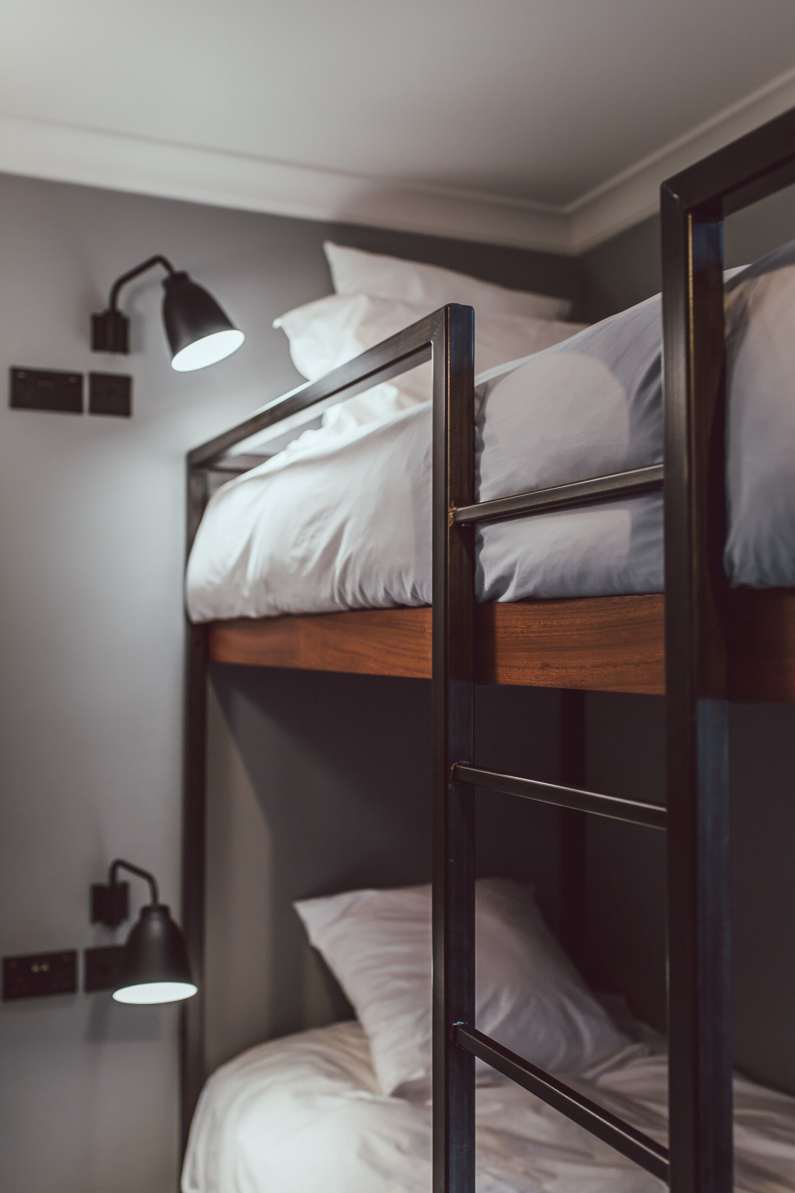 A bunk bed with two levels, each with white bedding and a black metal frame. There are two wall-mounted lamps providing light.