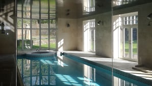 Una piscina interior con paredes de vidrio transparente, suelo de baldosas y vistas a un jardín exterior.