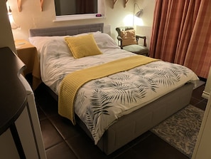 A neatly made bed with a tropical leaf pattern bedspread, a yellow quilted throw, and a wooden nightstand with a lamp.