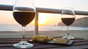 Two wine glasses on a wooden table with cheese and crackers, overlooking a sunset and the ocean. - Alojamento Batata - Peixe Imperador  (Nazaré)