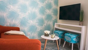 A room with a patterned wall, a sofa, a small round table with decorative items, and a TV on a white stand. - Alojamento Batata - Peixe Imperador  (Nazaré)