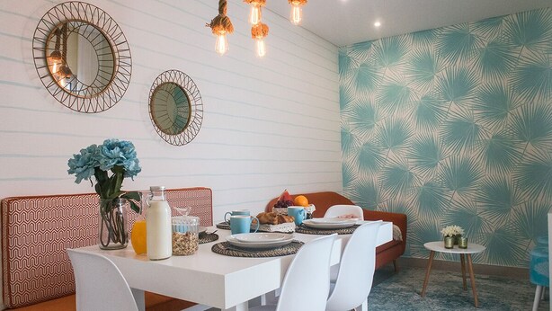 A dining area with a white table, chairs, and a patterned wall. There are two wall mirrors, a hanging light fixture, and a small round table with a vase and a fruit bowl. - Alojamento Batata - Peixe Imperador  (Nazaré)