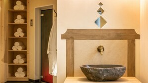 Un baño con lavamanos de piedra, estantes de madera con toallas y un espejo con decoración geométrica en la pared.