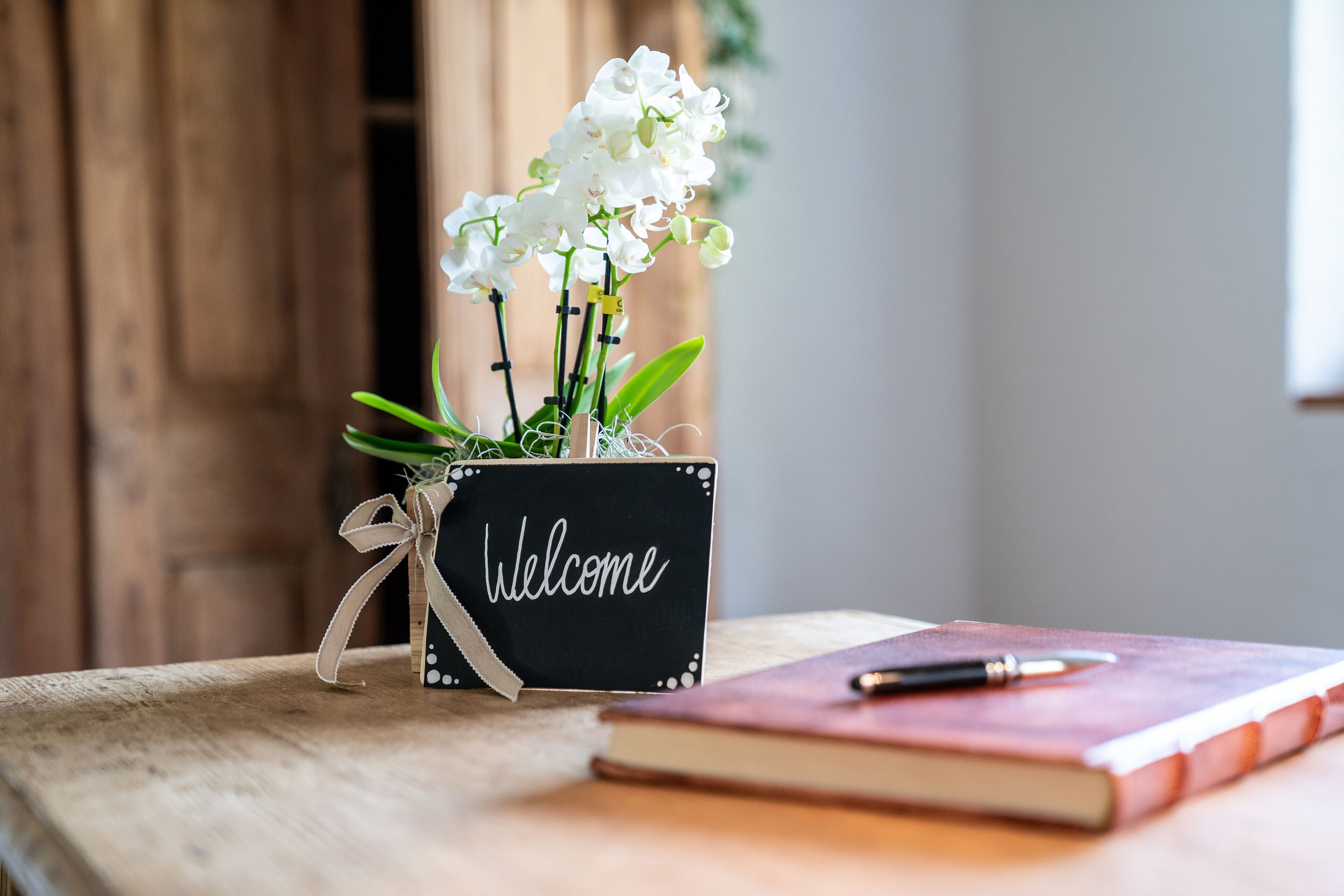A wooden table with a black box containing white flowers and a 'Welcome' sign, and a notebook with a pen on it.
