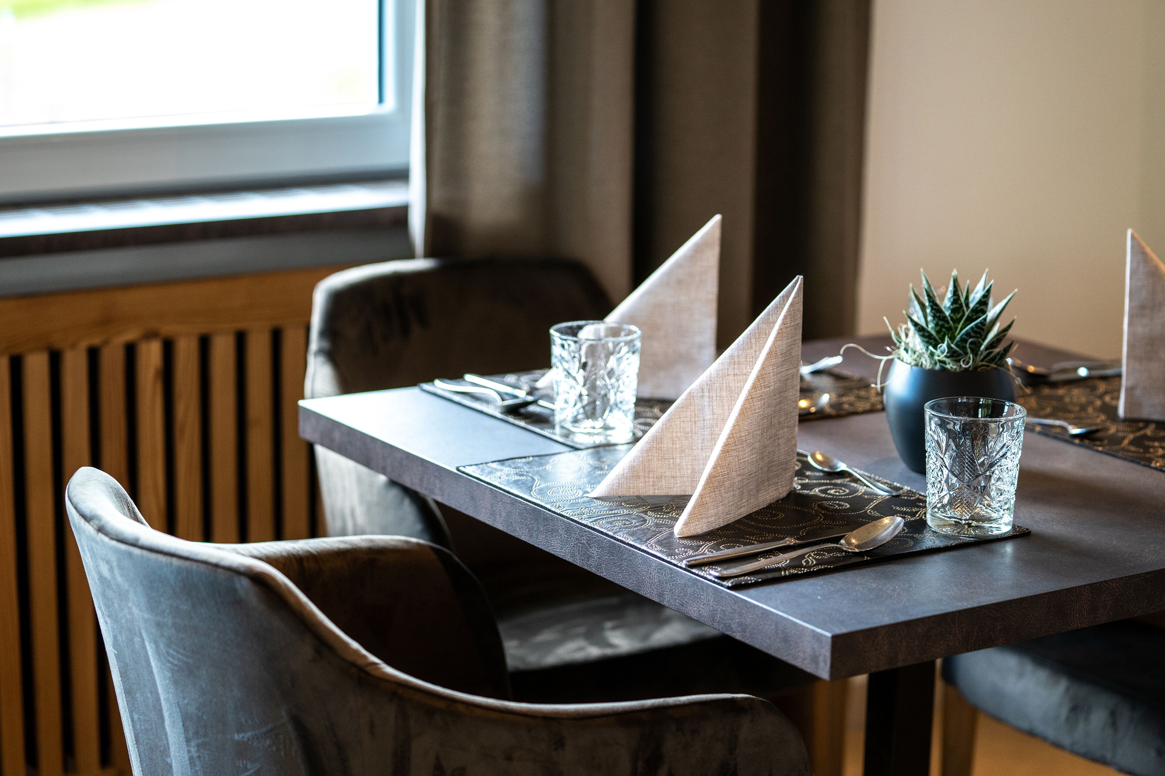 Una mesa de comedor dispuesta con un mantel individual, servilletas, cubiertos, una copa y una pequeña planta en maceta.