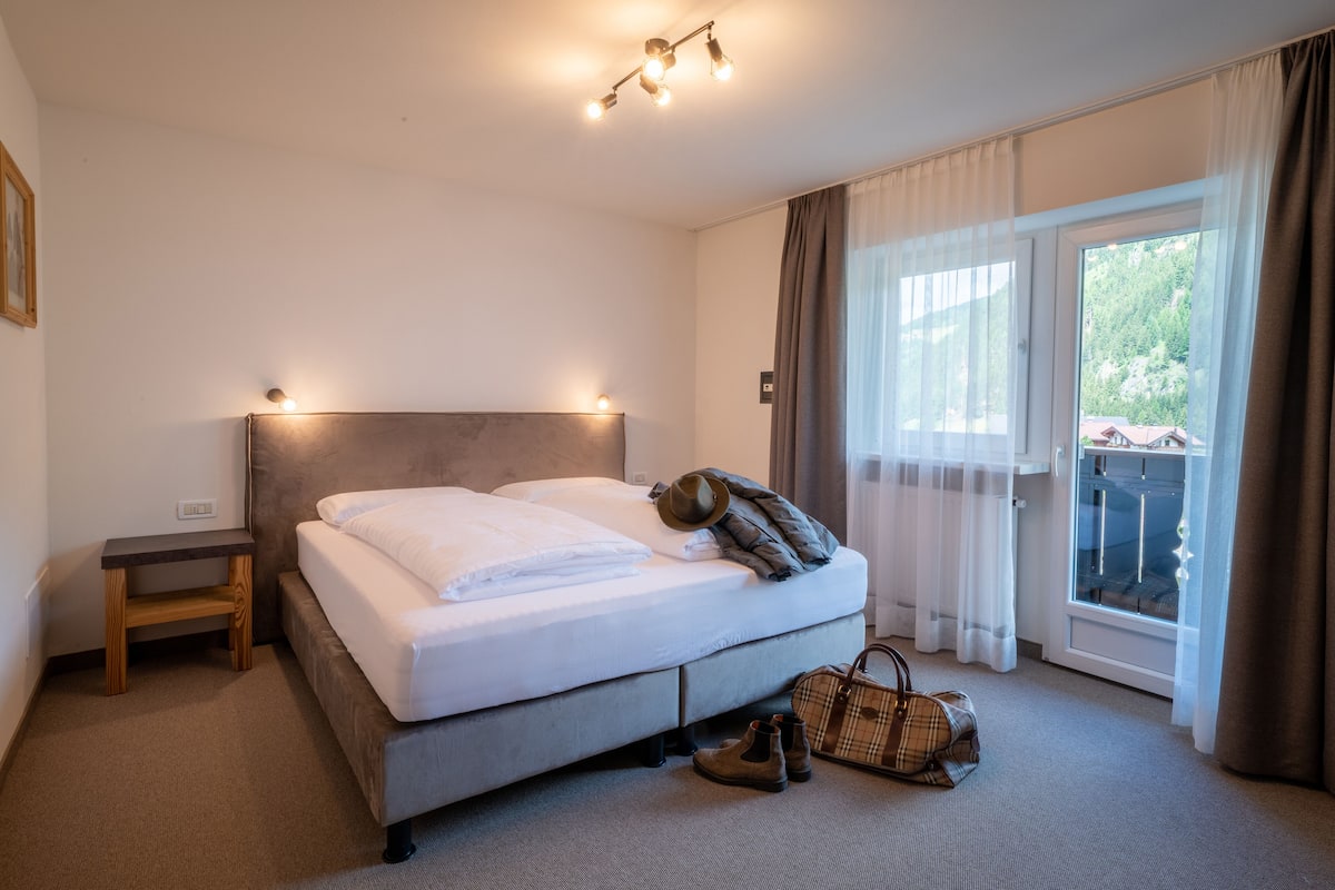 A neatly made bed with a headboard, a small wooden nightstand, a pair of boots, and a handbag on the floor.