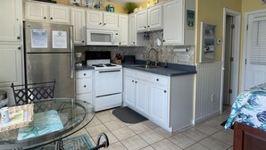 A compact kitchen with white cabinets, a stainless steel refrigerator, a microwave, and a stove. There is a small dining area with a glass table and chairs. - Come and Relax at Paradise Found! (HATTERAS)