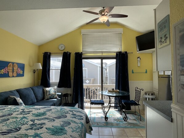 A bedroom with a bed, a ceiling fan, a window with curtains, a small table, and a chair.