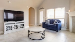 A living room with a flat-screen TV on a white cabinet, a round coffee table, a blue sofa with patterned cushions, and a white fireplace.