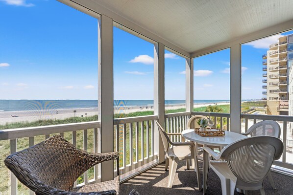 A balcony with a table and chairs, overlooking a beach and a building. - Dunescape 201-3BR/2.5BA Oceanfront condo in Crescent Beach (North Myrtle Beach)