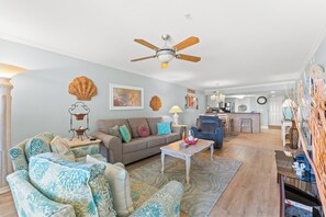 A living room with a sofa, armchairs, a coffee table, a ceiling fan, and a kitchen in the background.