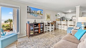 A modern living room with a sofa, a coffee table, and a TV. There is a dining area with white chairs and a kitchen with white cabinets.