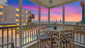 A balcony with a view of the ocean and a sunset. There is a table with chairs and a ceiling fan.