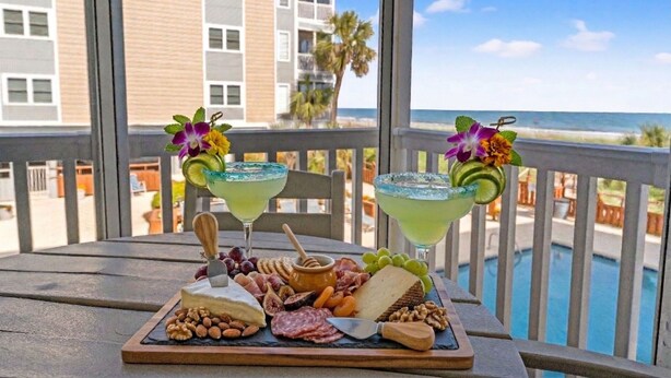 A table with a charcuterie board, two cocktails with decorative flowers, and a view of a pool and beach.