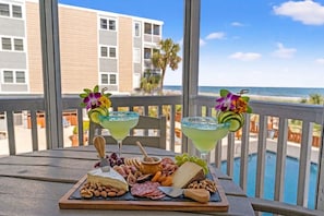 A table with a charcuterie board, two cocktails with decorative flowers, and a view of a pool and beach. - Palmwood Villas 104-2BR/2BA Oceanfront condo in Crescent Beach (North Myrtle Beach)