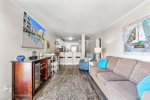 A living room with a sofa, a coffee table, a rug, a painting, and a view into the kitchen. - Palmwood Villas 104-2BR/2BA Oceanfront condo in Crescent Beach (North Myrtle Beach)