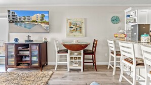 A modern kitchen with white cabinetry, a dining area with a wooden table and chairs, a television displaying a pool view, and a clock on the wall.