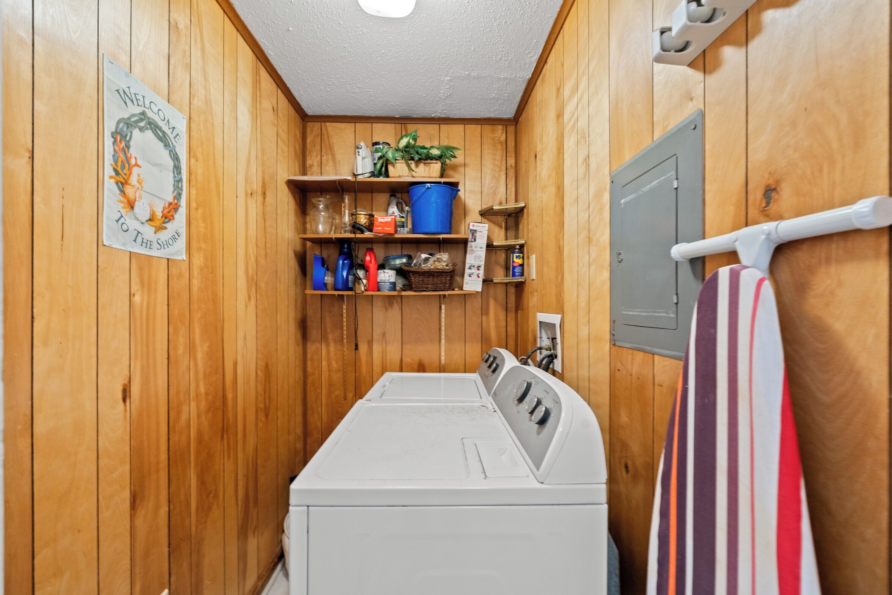 A laundry room with wooden walls, a washing machine, shelves with cleaning supplies, and a welcome sign.